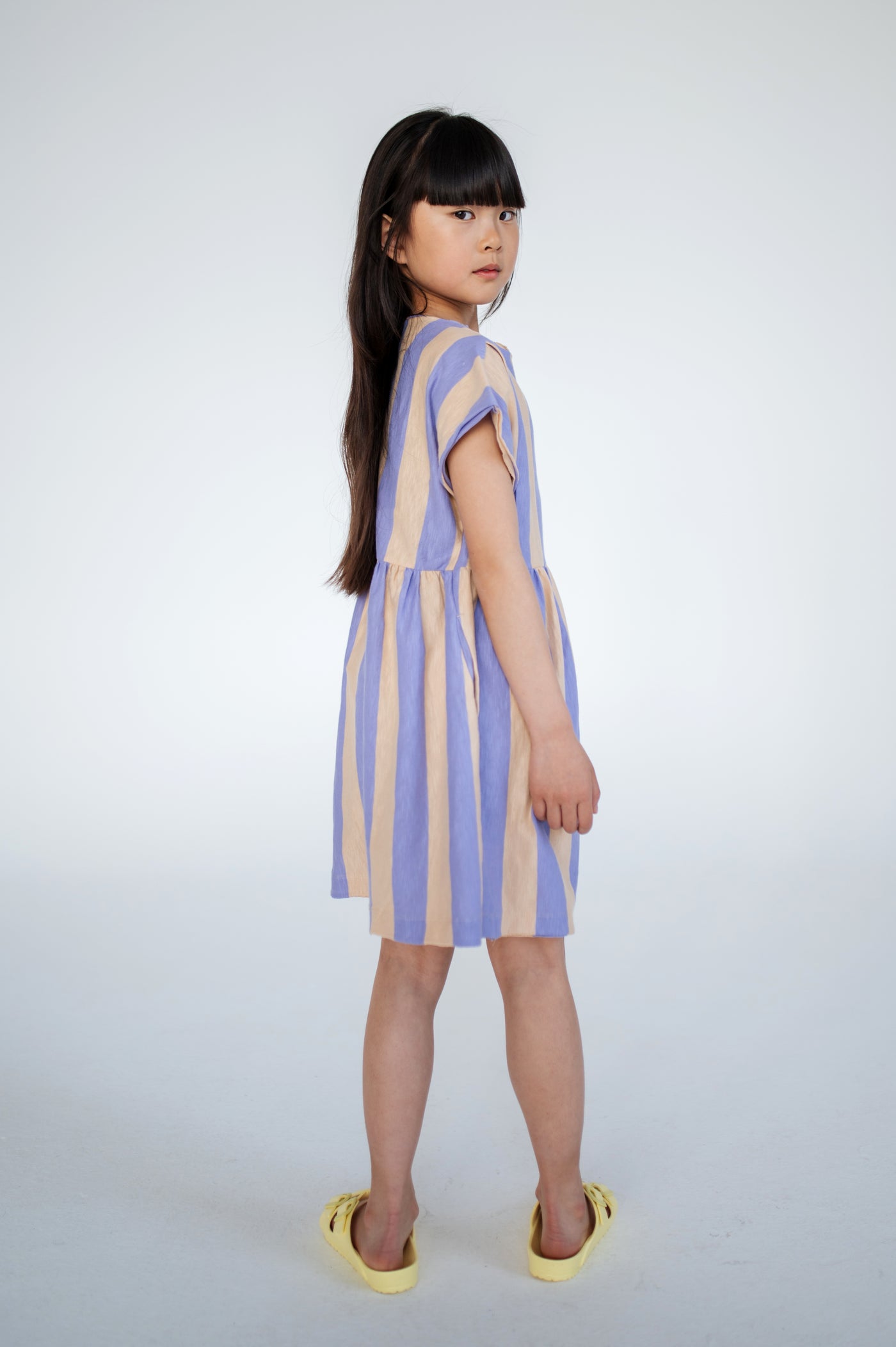 Lilac nude stripe dress made from 100% yarn-dyed organic cotton flamé, offering rich, lasting color and texture. Features pleated front details, ruffled waist, side pockets, short sleeves, and raw edge neckline. Lightweight and breathable for comfortable everyday wear.