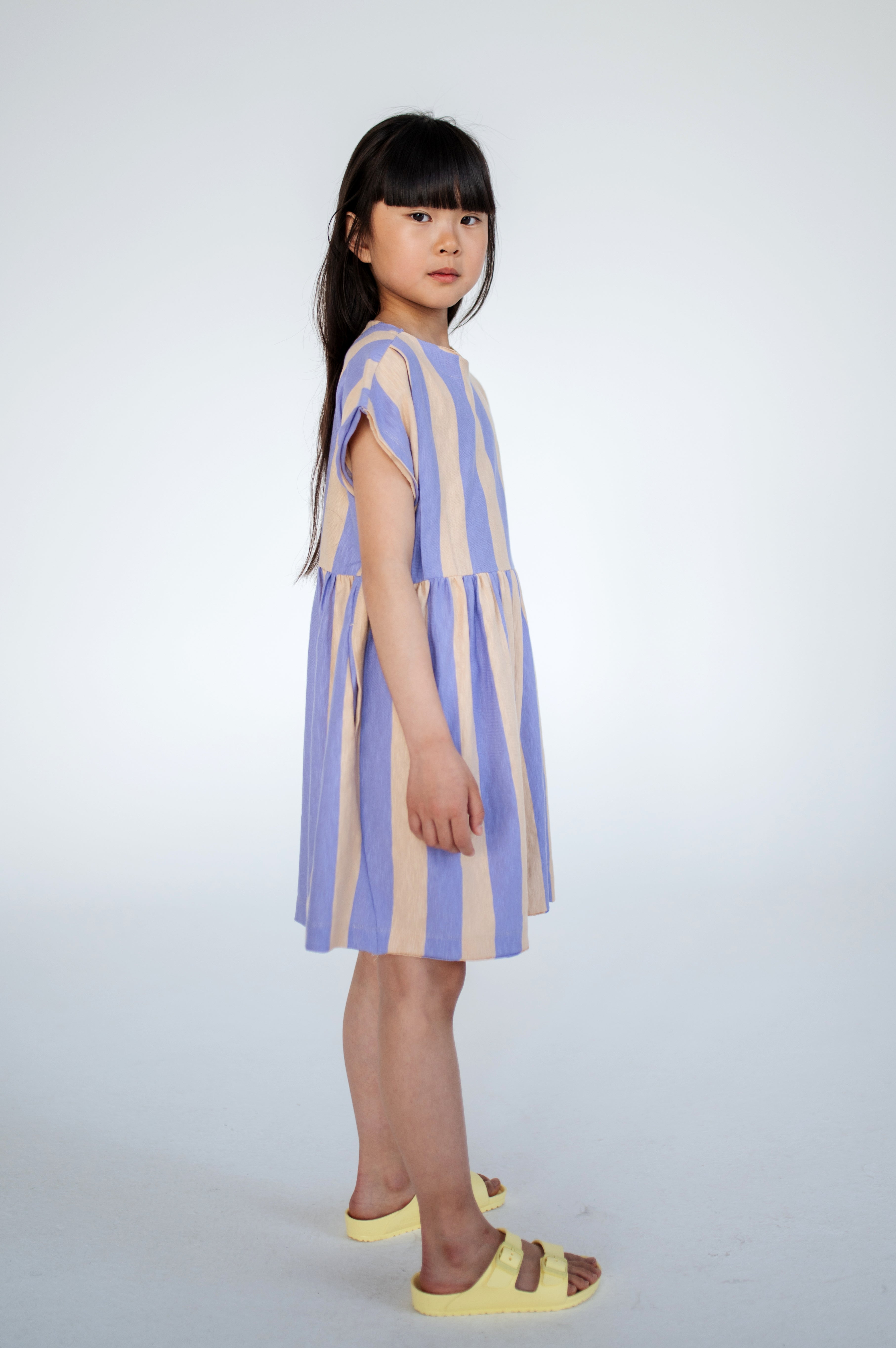 Lilac nude stripe dress made from 100% yarn-dyed organic cotton flamé, offering rich, lasting color and texture. Features pleated front details, ruffled waist, side pockets, short sleeves, and raw edge neckline. Lightweight and breathable for comfortable everyday wear.