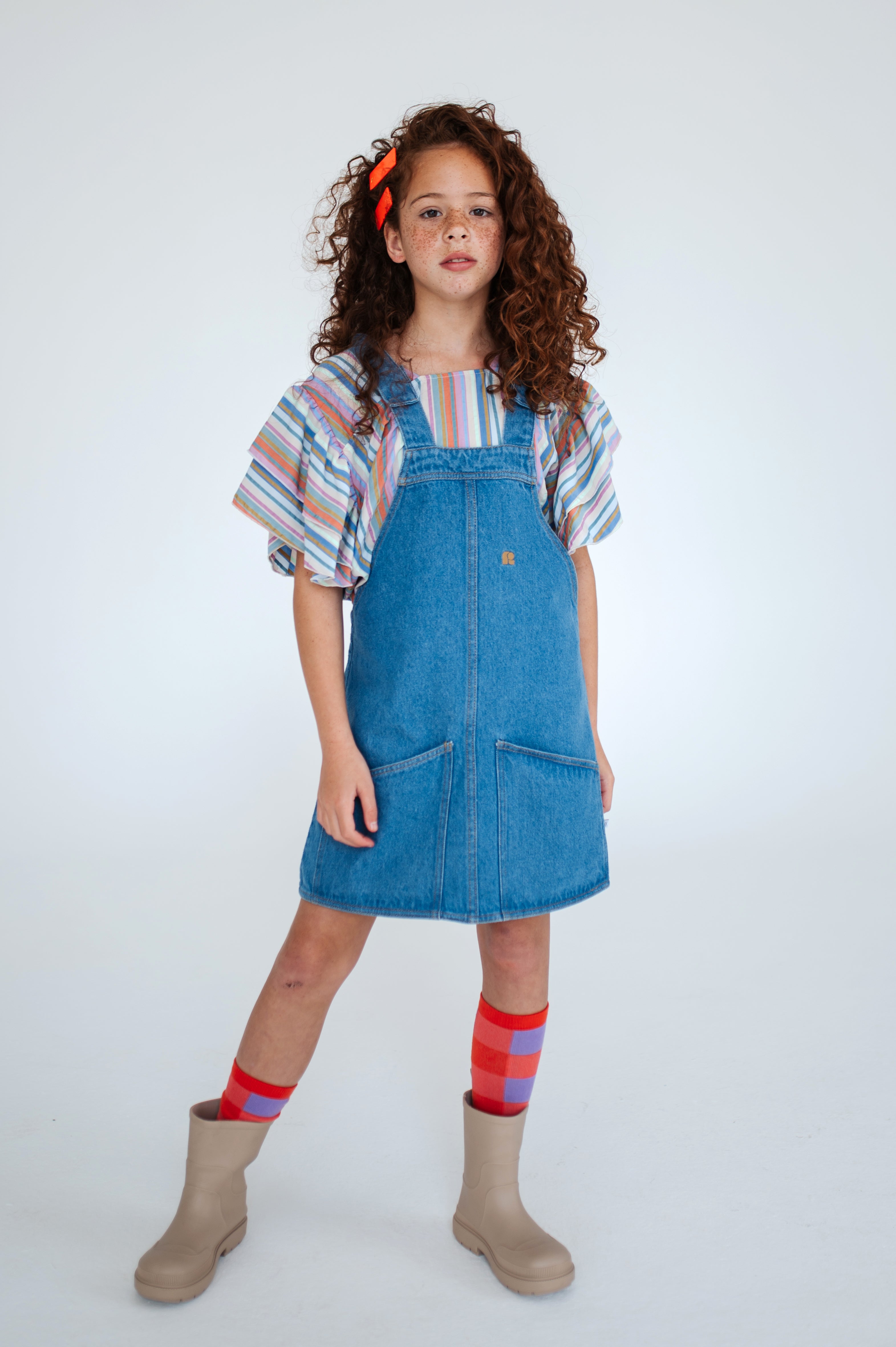 Medium blue dungaree dress made from durable organic cotton denim. Features adjustable snap-button shoulder straps, front and back patch pockets, A-line skirt, embroidered logo, and woven brand tag.