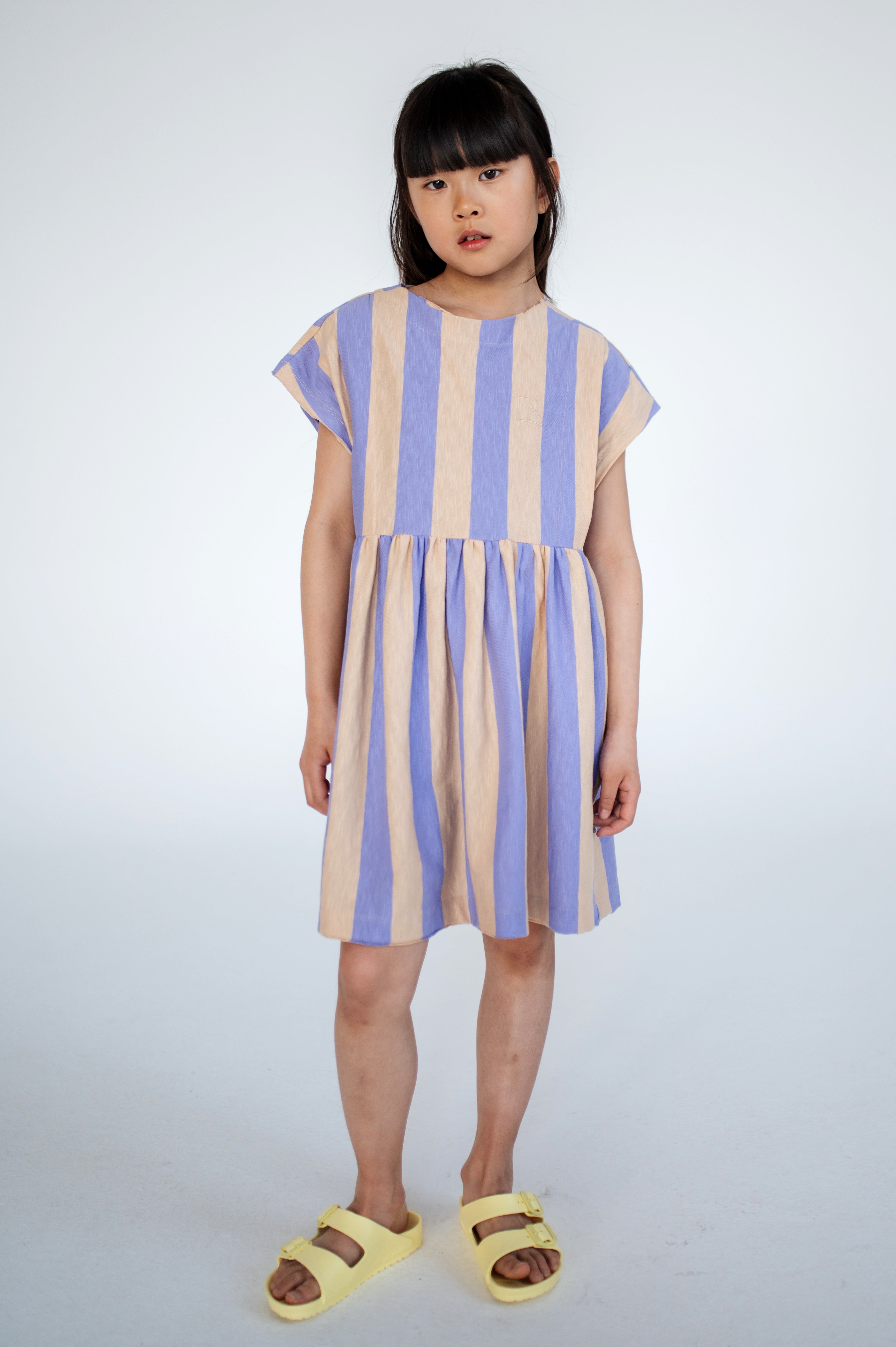 Lilac nude stripe dress made from 100% yarn-dyed organic cotton flamé, offering rich, lasting color and texture. Features pleated front details, ruffled waist, side pockets, short sleeves, and raw edge neckline. Lightweight and breathable for comfortable everyday wear.